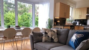 Una sala de estar moderna con un sofá gris, un comedor con sillas blancas y una cocina con armarios de madera.