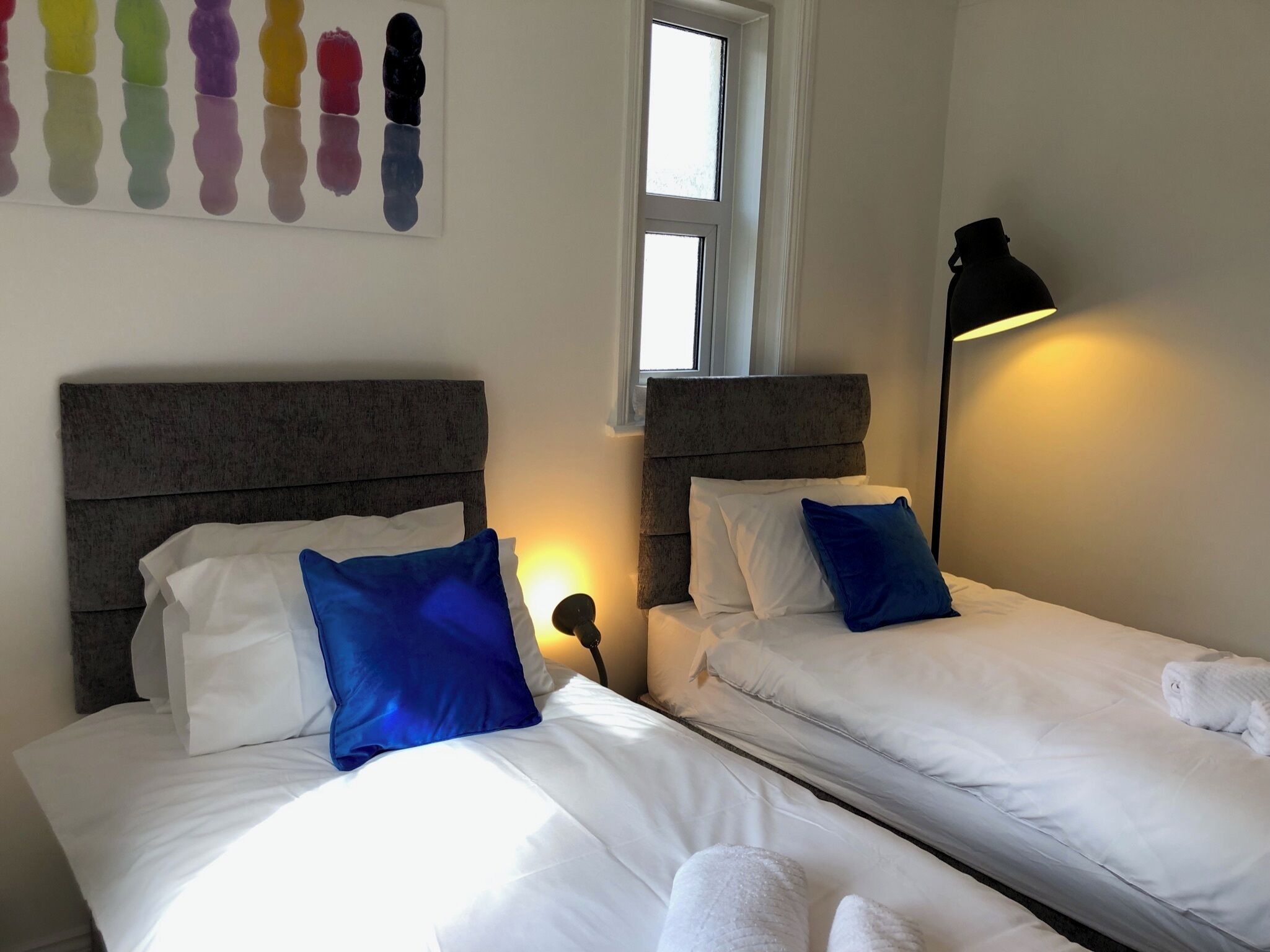 A hotel room with two beds, each with white bedding and blue pillows, a window, and a bedside lamp.