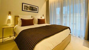Habitació d'hotel amb un llit gran, dues taules de nit amb làmpades, un quadre amb marc a la paret i una finestra gran amb cortines translúcides.