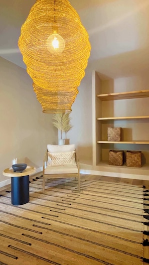 A room with a large, textured hanging light fixture, a white armchair, a small round table, a patterned rug, and a bookshelf with decorative items. - The Cliffs - PradaBeach (Xove)