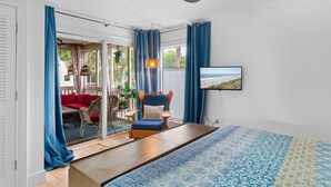 A room with a ceiling fan, a TV on the wall, a wooden table, a blue patterned rug, and a view of the outdoors through a sliding door.