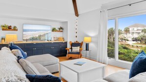 A living room with a sofa, coffee table, and a chair with a sign. There is a kitchen area with cabinets and a TV. A large window offers a view of a house and greenery.