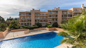 Une piscine entourée d’une clôture blanche, bordée de palmiers et surplombée d’un immeuble à plusieurs étages doté de balcons.