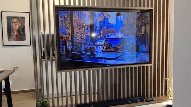 En moderne stue med et stort fladskærms-tv monteret på en væg af træpaneler, et indrammet kunstværk til venstre, og et lille bord med en bærbar computer og en kop.