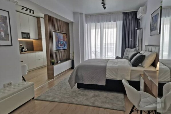 A modern hotel room with a bed, desk, and kitchenette.