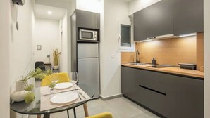 A compact kitchen with modern appliances, a dining area with yellow chairs, and a small refrigerator. - Home in Athens (Kallithea)