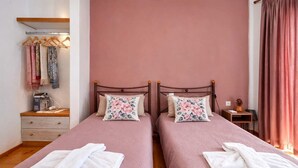 A twin bedroom with two beds, each with a floral pillow, a wooden nightstand, a small table, and a hanging rack with clothes. - Dinas Apartment Alonissos - Nefeli Summer Haven (Patitiri)