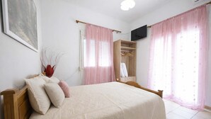 A bedroom with a bed, a wooden wardrobe, a TV mounted on the wall, and pink curtains. - Flower's Studio Alonnisos - Artemis Summer Escape (Patitiri)