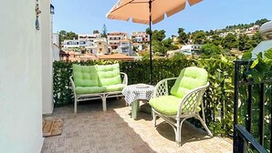 Pequeño patio al aire libre con dos sillas verdes con cojines y una mesa, un paraguas color beige y vistas a un pueblo en la ladera.