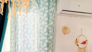 Una habitación con una cortina de hojas azules y blancas, una mesita de noche pequeña con un objeto decorativo y un aire acondicionado instalado en la pared.