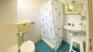 Un baño con inodoro, lavabo, cortina de ducha con motivos de delfines y un taburete pequeño.