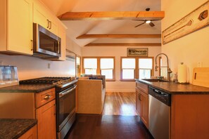 Une cuisine avec des meubles en bois, un four à micro-ondes, une cuisinière et un lave-vaisselle. Il y a des fenêtres avec des stores et un ventilateur de plafond.