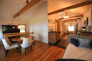 Uma sala de estar moderna com uma mesa de jantar de madeira, duas cadeiras, um sofá, uma área de cozinha com pia e fogão, e uma televisão.