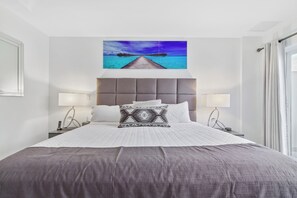 Una cama bien hecha con un cojín estampado, dos lámparas de noche y un cuadro en la pared que muestra un muelle sobre el agua.