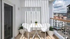 Un balcón con una mesa y sillas blancas, plantas en macetas y vistas de la ciudad.