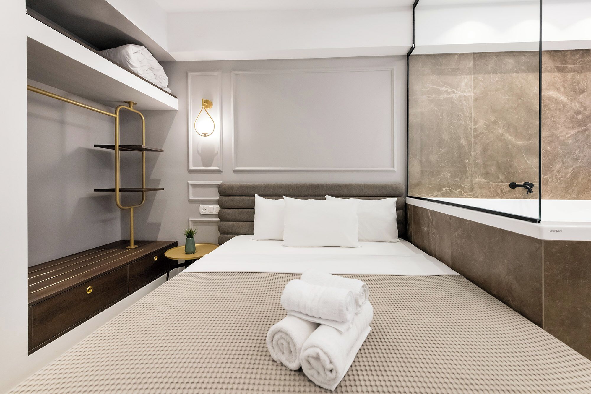 Une chambre d’hôtel moderne comprenant un lit, une étagère intégrée, une table de chevet, une petite table ronde et un grand miroir.
