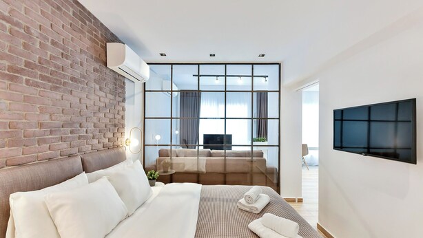 Una habitación de hotel moderna con una pared de ladrillo decorativa, una cama grande con ropa blanca, un televisor de pantalla plana montado en la pared y un ventanal con vistas a la ciudad.