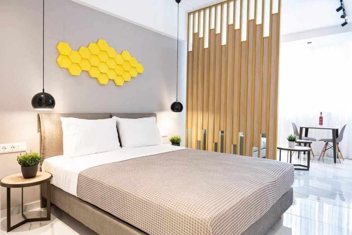 Une chambre moderne avec un grand lit, un luminaire jaune en forme d’hexagone fixé au mur, une cloison en lattes de bois et une petite table avec des chaises.