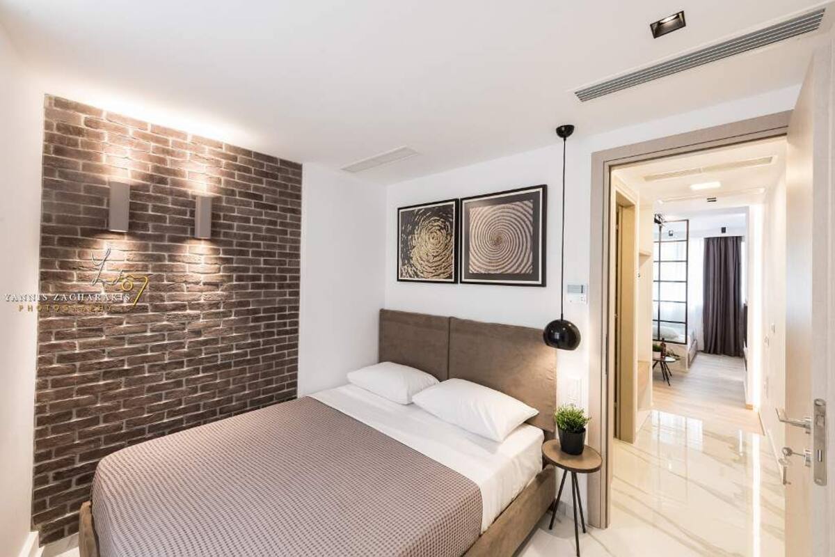 Une chambre moderne avec un mur d’accent en briques, un lit recouvert de linge blanc, deux œuvres d’art encadrées et une petite table d’appoint agrémentée d’une plante.
