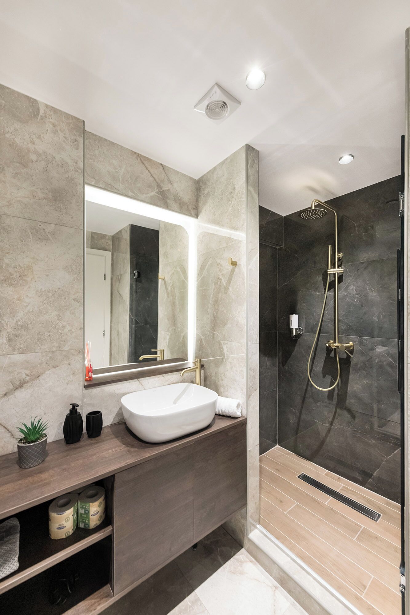 A modern bathroom with a large mirror, a white sink, a shower with a gold showerhead, and dark marble walls.