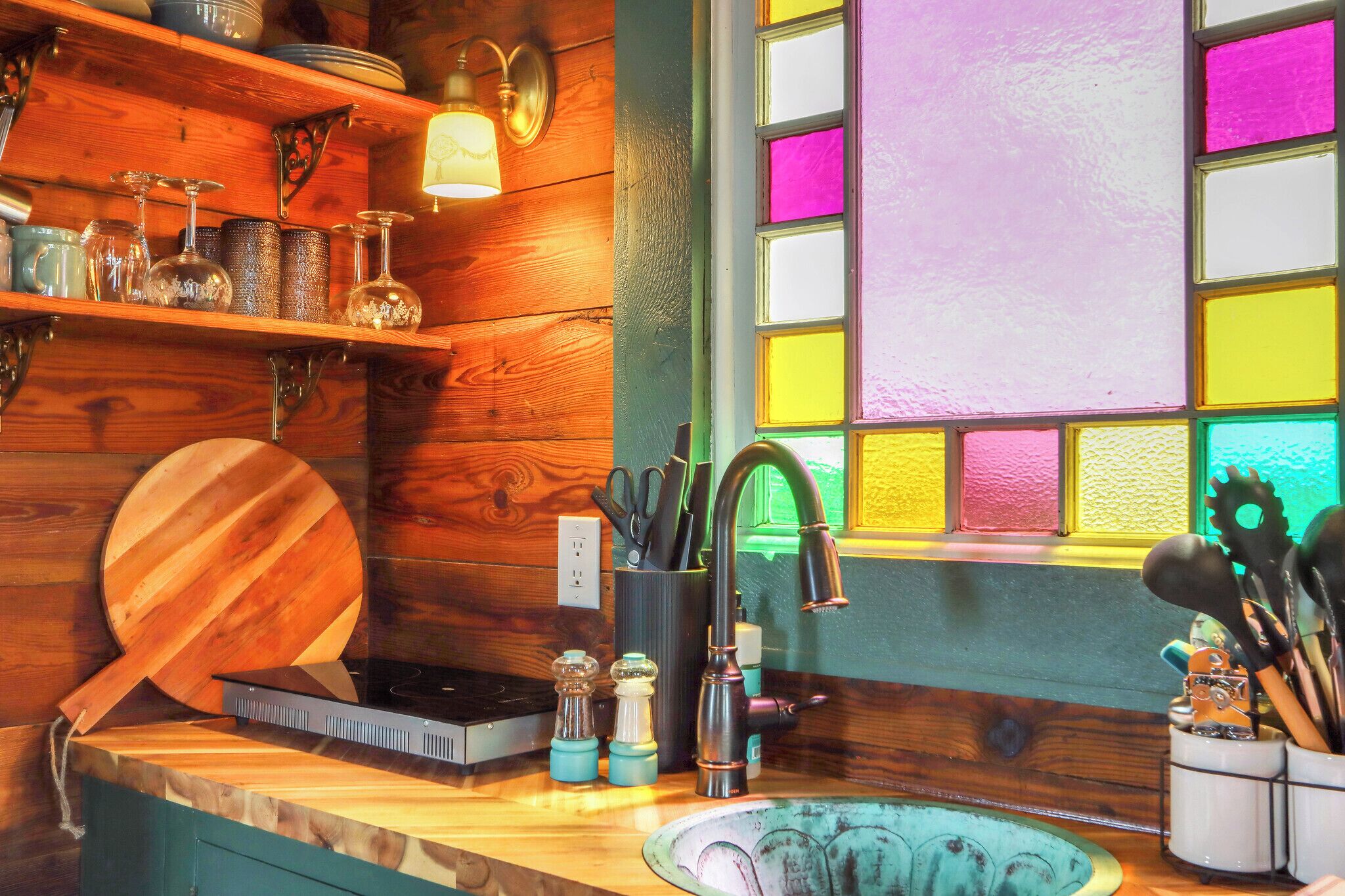 Una cocina con estantes de madera, una tabla de cortar, un fregadero y un ventanal con vidrios de colores.