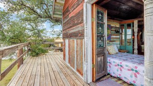 A rustic wooden cabin with a wooden deck, a bed with a quilt, and a glass door leading to the interior. - Romantic Treehouse Escape in Hill Country (Lampasas)