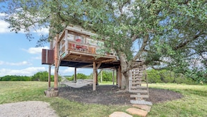 A treehouse with a wooden deck and a ladder, surrounded by a grassy area and a dirt path. - Romantic Treehouse Escape in Hill Country (Lampasas)