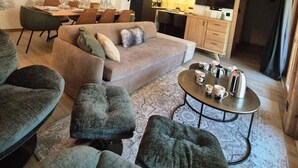 A modern living room with a sofa, ottomans, and a coffee table. There is a dining area with chairs and a table, and a kitchen in the background.