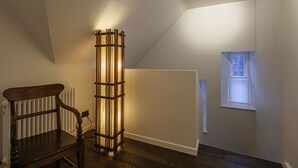 Una habitación con un radiador, una lámpara de pie alta, una silla de madera y un ventanal pequeño.