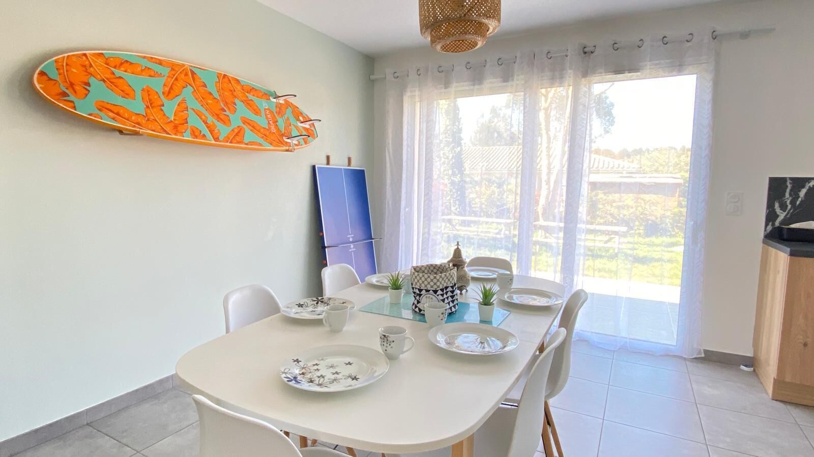 Un coin repas avec une table dressée pour quatre personnes, une planche de surf accrochée au mur et une vue sur l’extérieur grâce à de grandes fenêtres.