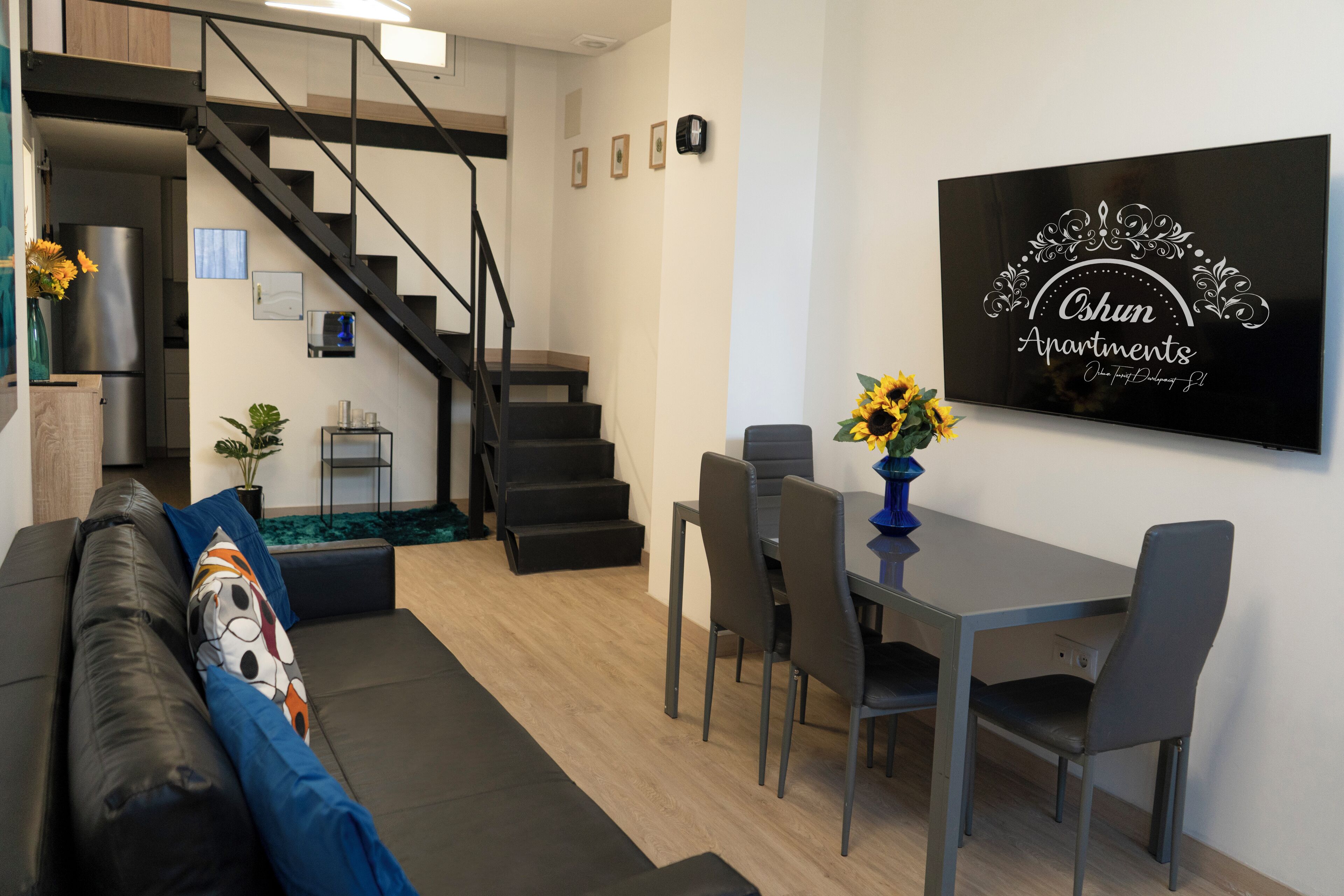 A modern living room with a sofa, a dining table with chairs, a staircase, and a wall-mounted sign.