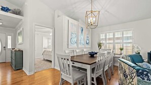 A dining area with a wooden table and white chairs, a chandelier, and artwork on the wall. - Steps Away-Golf Cart Deals-Fire Pit-Game Room (Destin)