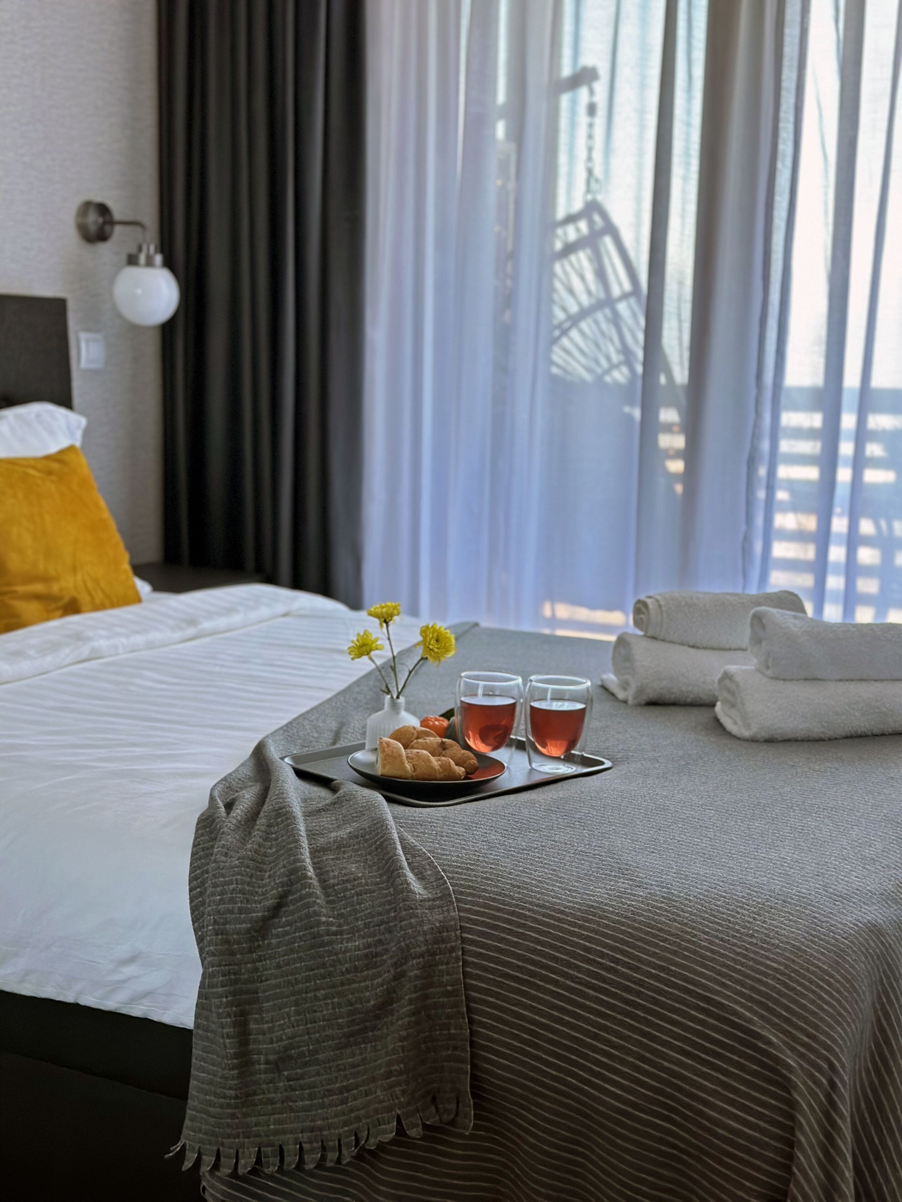 Un lit bien fait, recouvert d’une couverture grise, avec un oreiller jaune et un plateau contenant des croissants, deux verres de thé et un vase rempli de fleurs jaunes.
