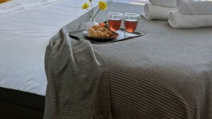 Un lit bien fait, recouvert d’une couverture grise, avec un oreiller jaune et un plateau contenant des croissants, deux verres de thé et un vase rempli de fleurs jaunes.