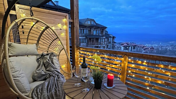 Un balcon avec une chaise en osier, une table ronde sur laquelle se trouvent une bouteille, des verres et une bougie, et une vue sur des bâtiments en arrière-plan.