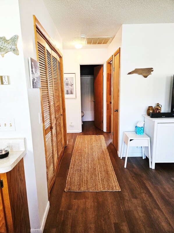 Un pasillo con piso de madera, una alfombra tejida, una pequeña mesa blanca con una caja de pañuelos y un cuadro en la pared.