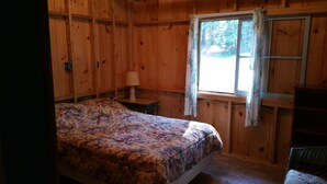 Une pièce de cabane en bois comprenant un lit, une table de chevet, une fenêtre avec des rideaux et une chaise.