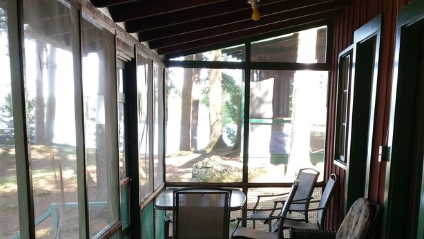 Un espace extérieur aménagé, abrité et doté d’un plancher en bois, de murs en verre avec une structure métallique, et offrant une vue sur une zone boisée.