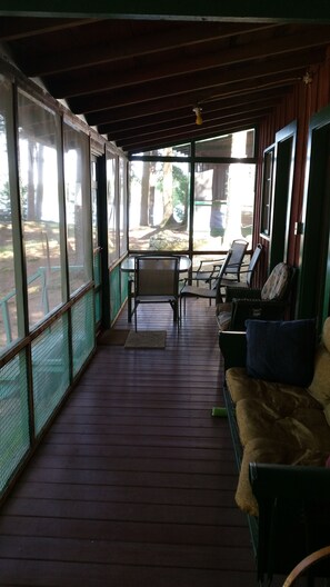 A covered outdoor seating area with wooden flooring, metal-framed glass walls, and a view of a forested area.