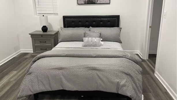Een slaapkamer met een bed, nachtkastje, lamp, een ingelijste foto en een raam.
