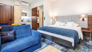Une chambre d’hôtel avec un canapé bleu, un lit doté d’une tête de lit grise et une table de chevet en bois.