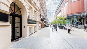 Une rue de la ville, avec des piétons, un immeuble portant une enseigne indiquant « APARTMENTY RUSSIAN », et un bâtiment en briques rouges en arrière-plan.
