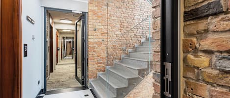 A modern interior with a staircase against a brick wall, a hallway with a geometric floor pattern, and a ceiling with a unique light fixture.