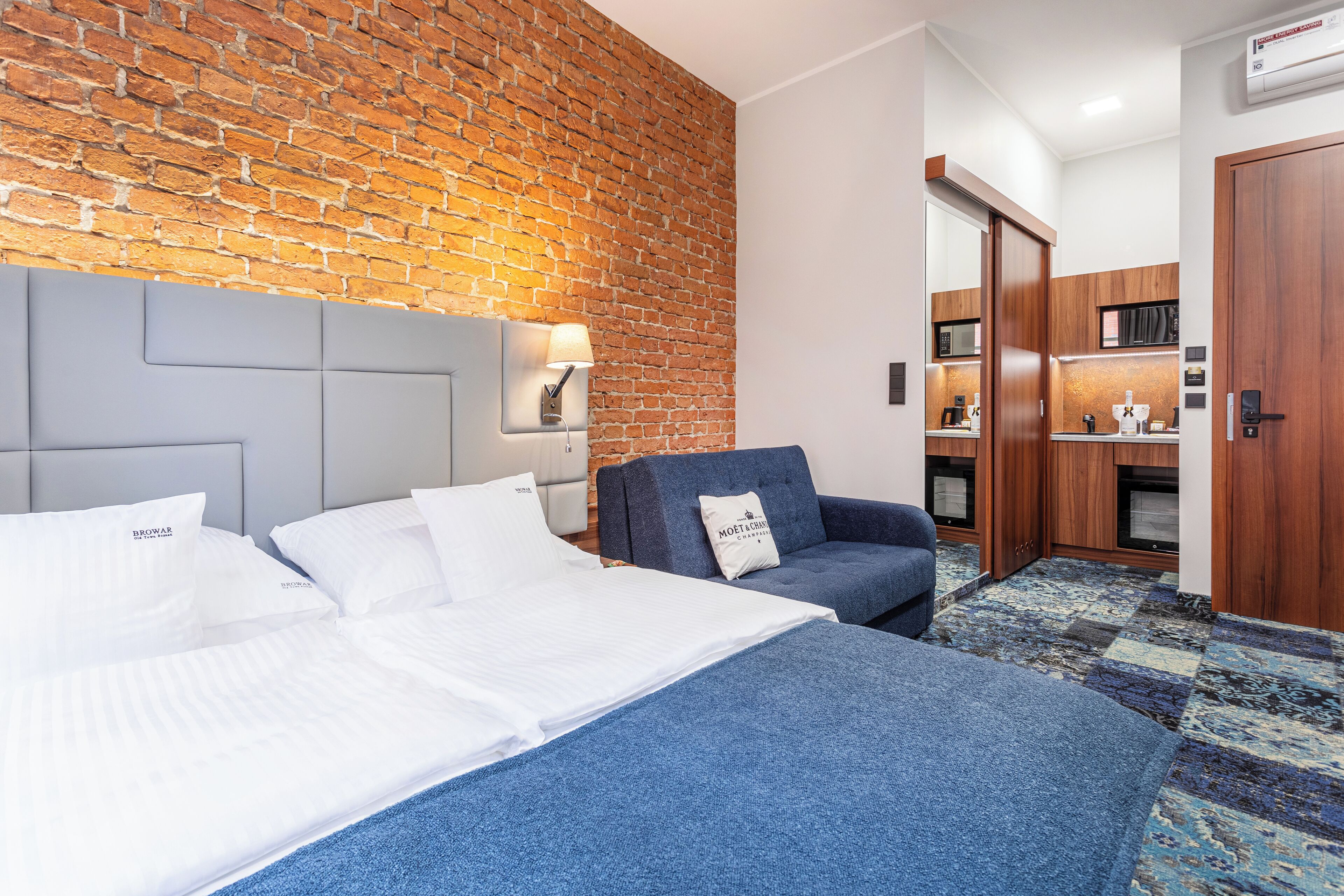 A hotel room with a brick wall, a bed with white bedding, a blue armchair, and a small kitchenette.