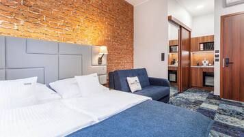 A hotel room with a brick wall, a bed with white bedding, a blue armchair, and a small kitchenette.