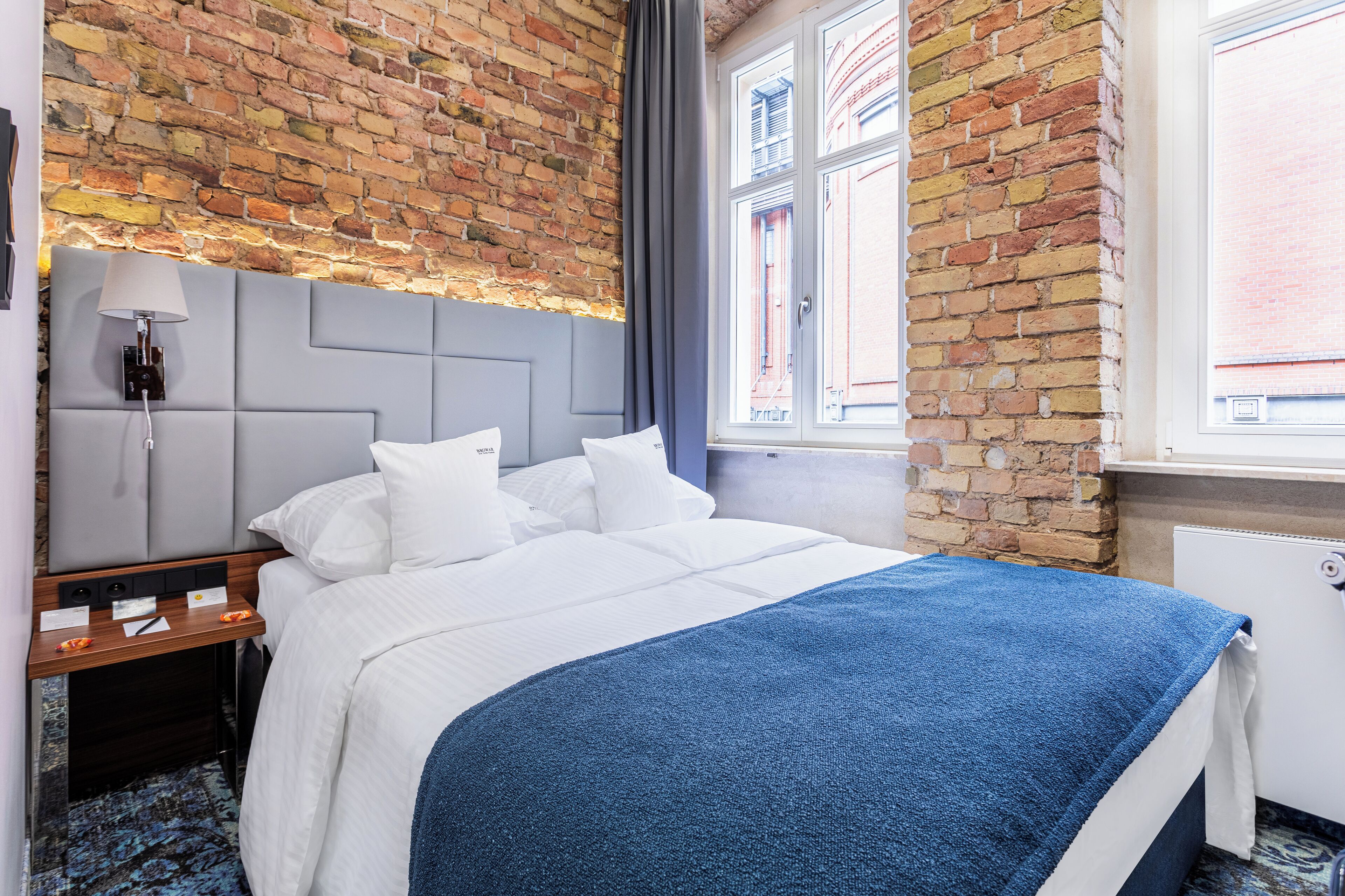 A hotel room with a brick wall, a large bed with white bedding, a blue throw blanket, a nightstand with a lamp, and a window with a view of buildings.