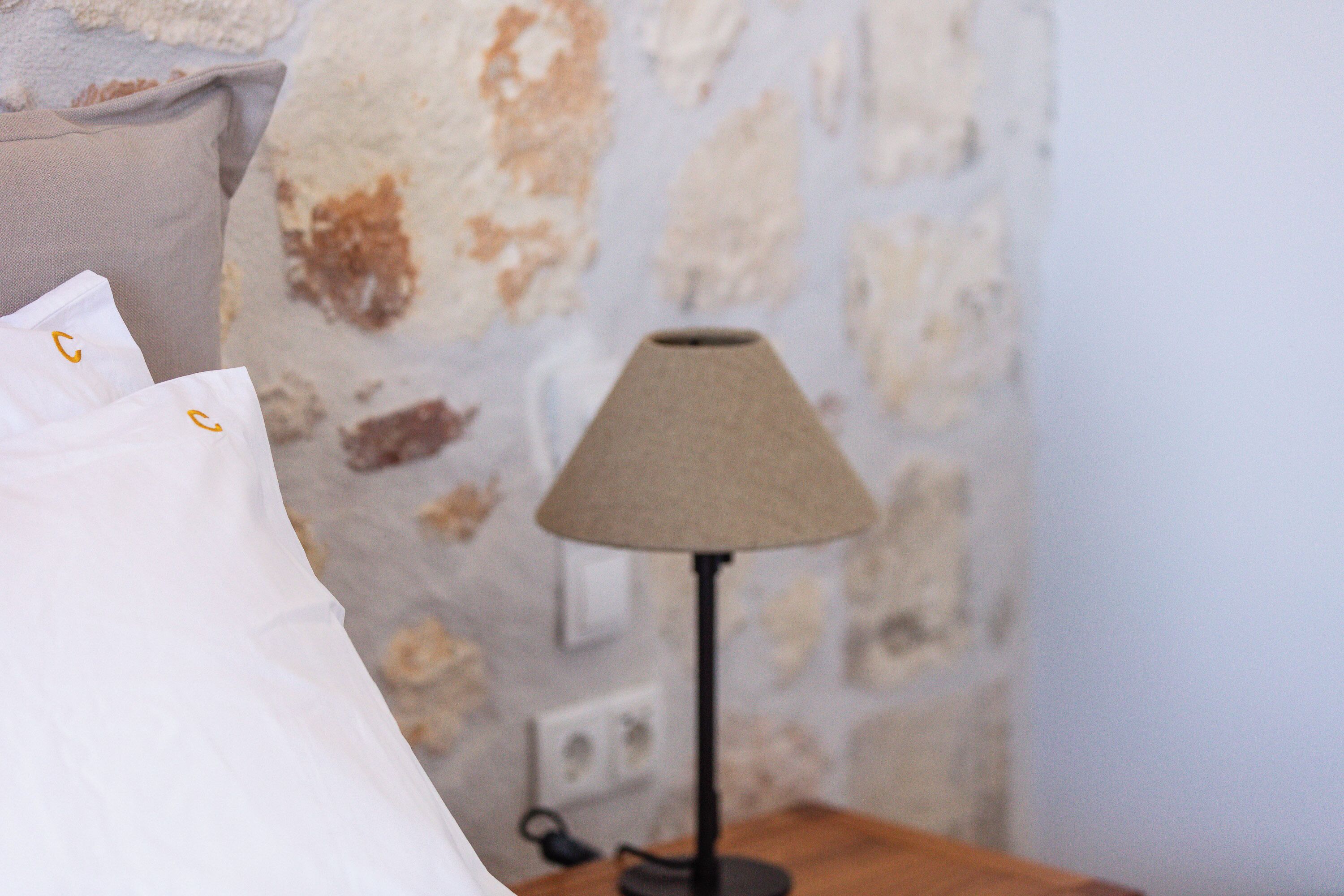 A bed with white linens and a lamp on a wooden nightstand against a stone wall.