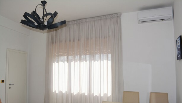 Una habitación con ventilador de techo, aire acondicionado blanco, un ventanal con cortinas translúcidas, sofá color beige y una pequeña mesa blanca.