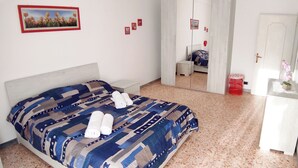 Una cama bien hecha con una colcha estampada y dos toallas enrolladas, una mesita de noche con un florero, y un espejo que refleja un dormitorio con cama y puerta.