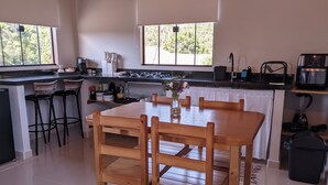 A modern kitchen with a wooden dining table and chairs, a built-in stove, and a large window with a view of trees. - Refuge in the mountains/near the trail to Pico do Papagaio Garcias Peak and Waterfall (Aiuruoca)